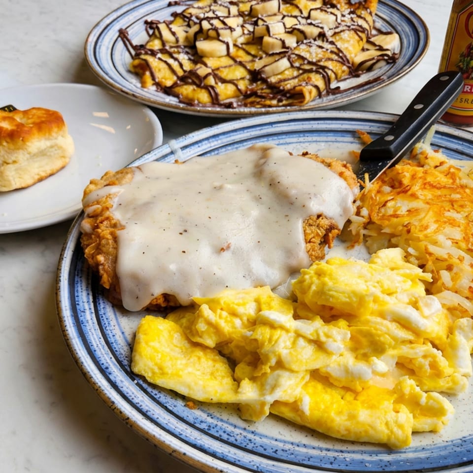Breakfast plate with scrambled eggs, hash browns, biscuits and gravy, alongside chocolate-drizzled pancakes and hot sauce.