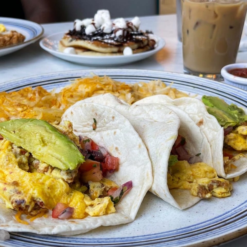 Breakfast plate with scrambled egg and avocado tacos, hash browns, salsa, iced coffee, and pancakes at a diner table.