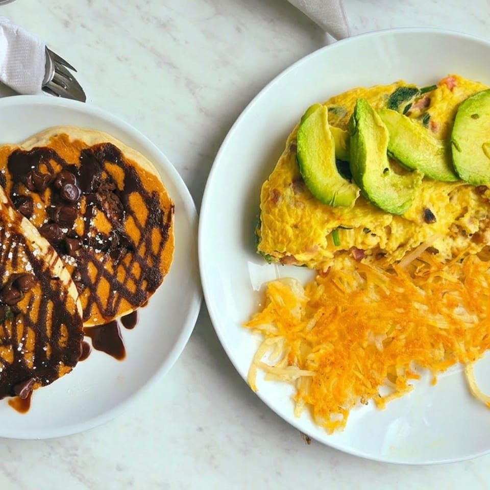 Breakfast spread with chocolate-drizzled pancakes, avocado-topped omelet, shredded hash browns, and orange juice on a marble table.